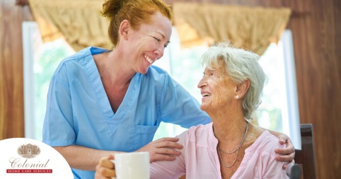 A caregiver and client smile while the caregiver hugs and helps the client, representing professional caregiving.